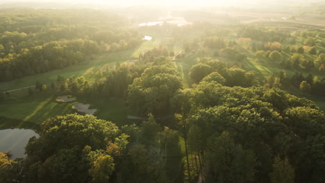 aerial view of golf course field at sunrise, green grassland lawn lands and trees in vast nature park forest in beautiful sunny autumn day, sunlight flare in horizon