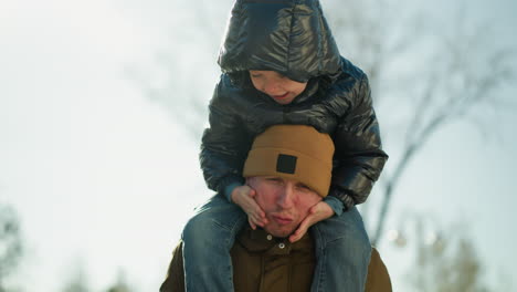 father carrying his son on his shoulders while the son playfully rubs his chin and laughs, the father appears to be enjoying the moment, with a soft light shining behind