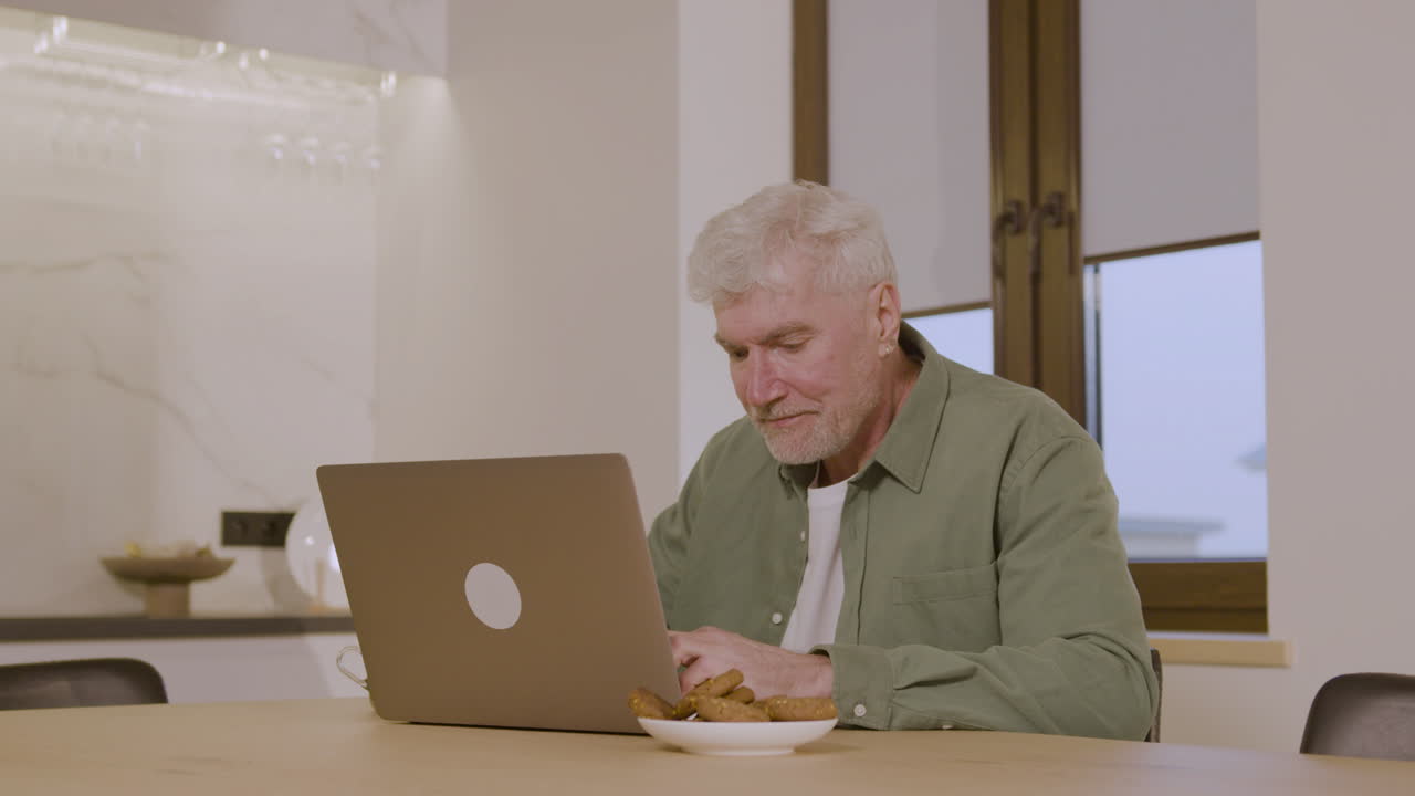 Happy Elderly Man Sitting On Chair In Kitchen And Using Laptop Computer