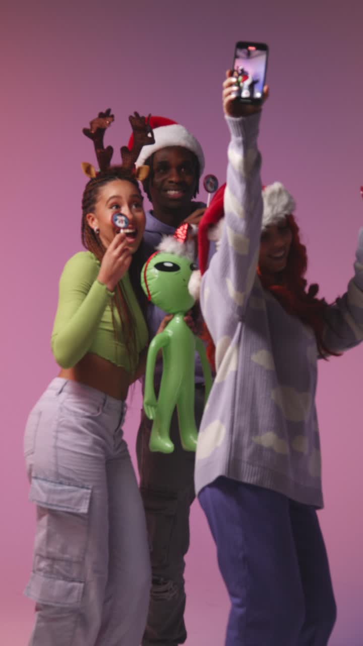 Vertical Video Studio Shot Of Gen Z Friends Dancing And Posing For Selfie At Christmas Party Wearing Santa Hat And Reindeer Antlers