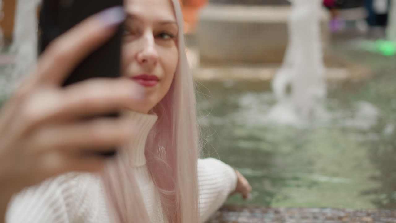 content creator touches fountain water while filming herself with phone near busy mall interior, capturing serene water play framed by blurred flowing fountain and modern shopping hall lights