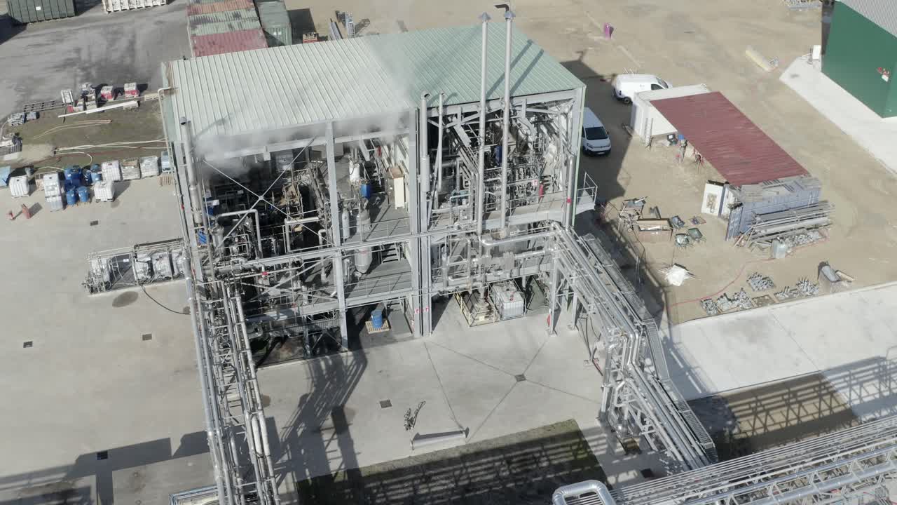 Drone footage showing exterior and some interior machinery of an open-structure industrial processing building with surrounding pipes, Castets, France