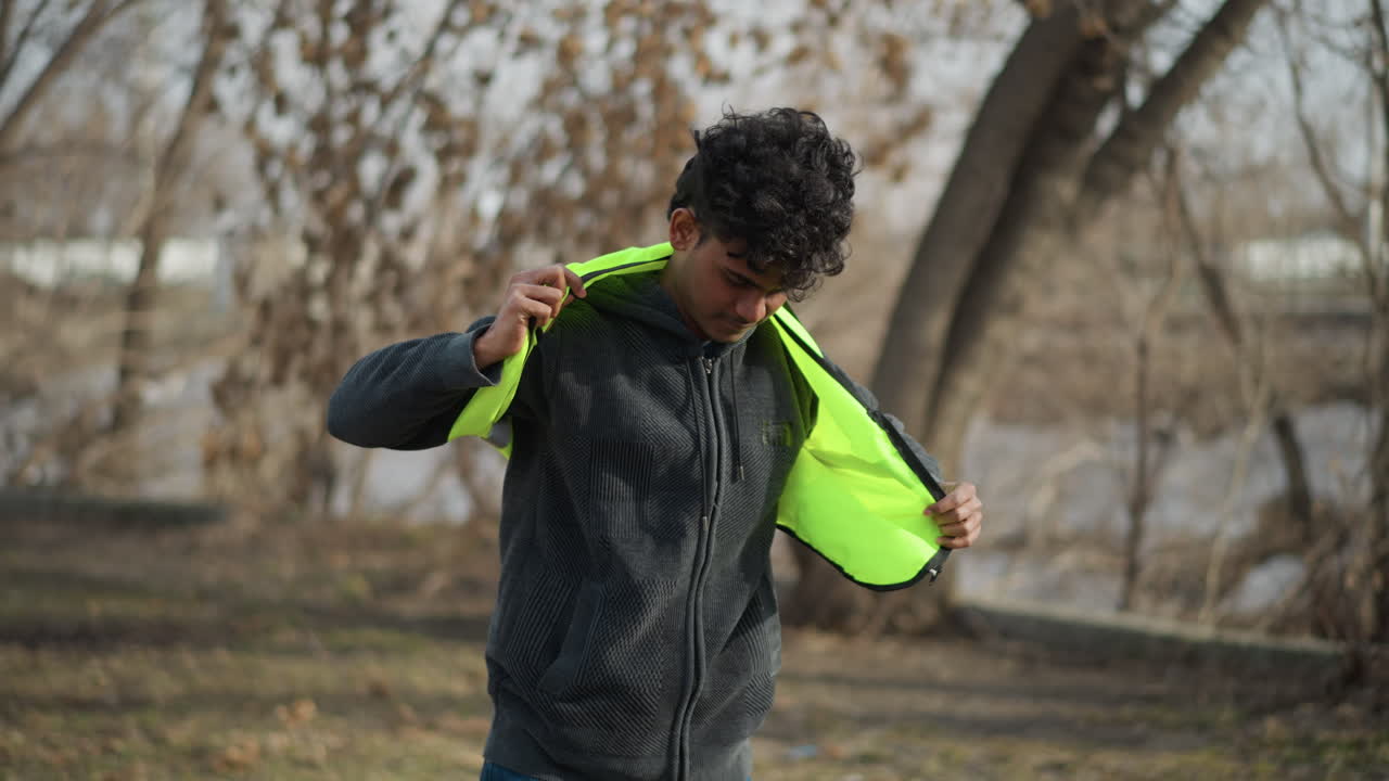 Man outdoors putting on bright neon green safety vest while standing in park with leafless trees, preparing for outdoor cleanup or volunteer work, backpack with gloves and water bottle placed on grass
