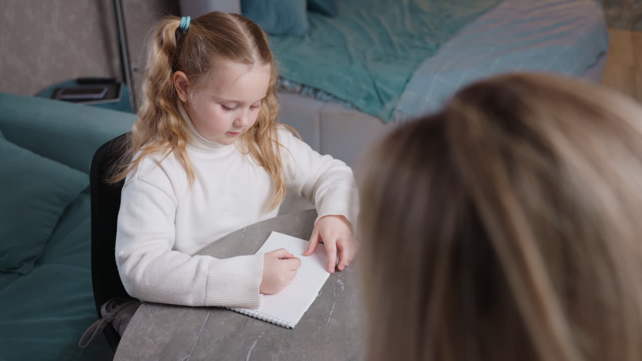 Niña rubia con jersey de cuello alto blanco sentada a la mesa entregando un cuaderno a una mujer durante una sesión de aprendizaje en casa, que simboliza la enseñanza, la cooperación, la educación, la crianza, el apoyo y la familia.