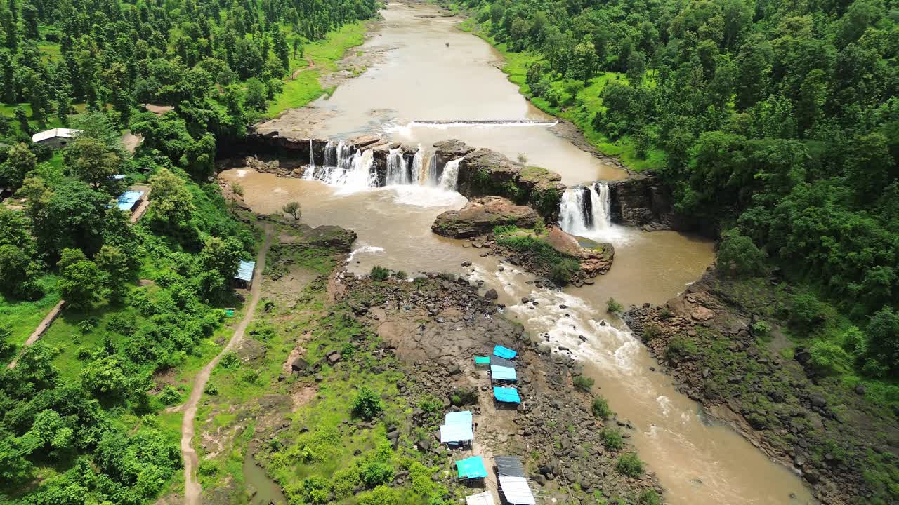 Aerial drone shot showing scenic multi tier waterfall flowing through rocky terrain surrounded by lush green forest, capturing flowing water, natural beauty, and peaceful landscape during daylight