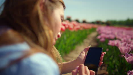 Back-view-of-young-woman-touching-smartphone-screen-with-fingers-in-flower-field