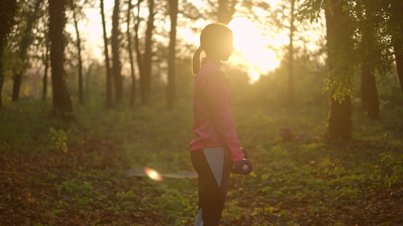 Woman Exercising with Dumbbell in Forest at Sunset