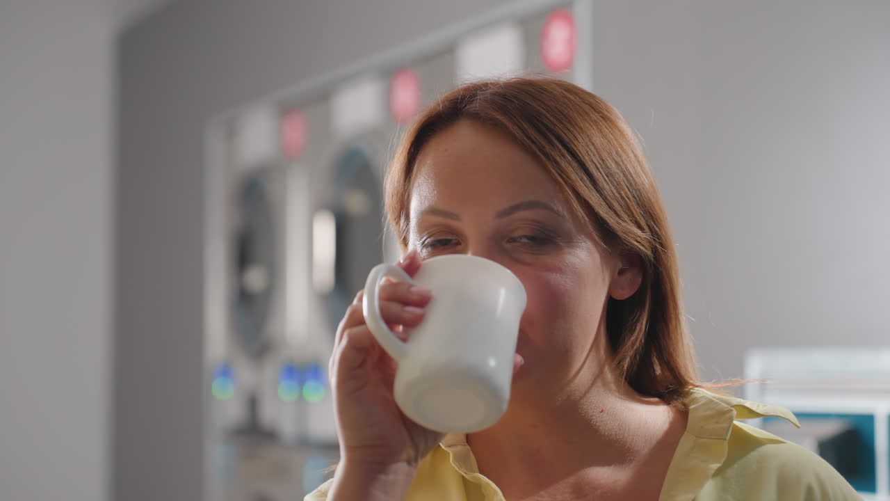 Portrait of laundry worker raising head, holding notebook, sipping coffee beside dryers and washers in modern laundromat, panels glowing, service pride, routine, professional cleanliness