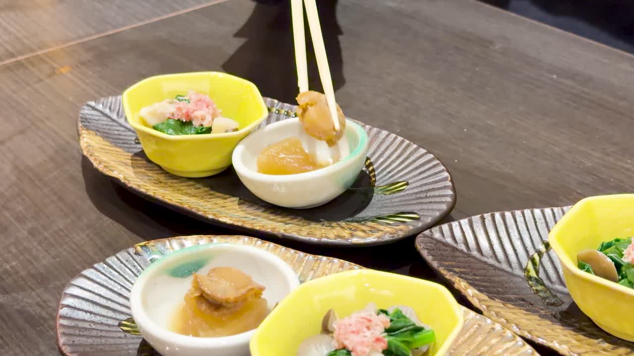 黄色い陶器の前菜ボウルからホタテと葉物野菜を箸でつまんでいるクローズアップショット。