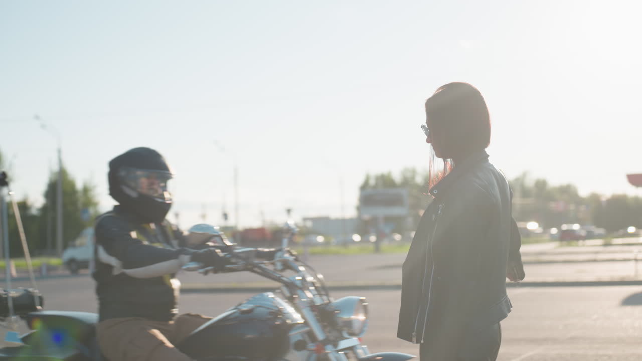 Confident woman in black leather jacket and sunglasses raises arm to stop rider under bright sunlight flare, standing on city street with bold attitude and modern urban lifestyle presence