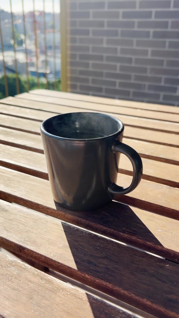 Aromatic morning coffee in a black ceramic mug sitting on a sunlit wooden slatted table on a balcony, with delicate steam rising from the hot beverage and casting a long shadow in the warm light