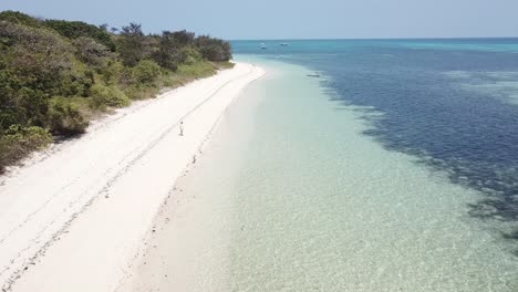 Drone-aerial-over-white-sandy-beach-with-someone-standing-by-the-blue-water