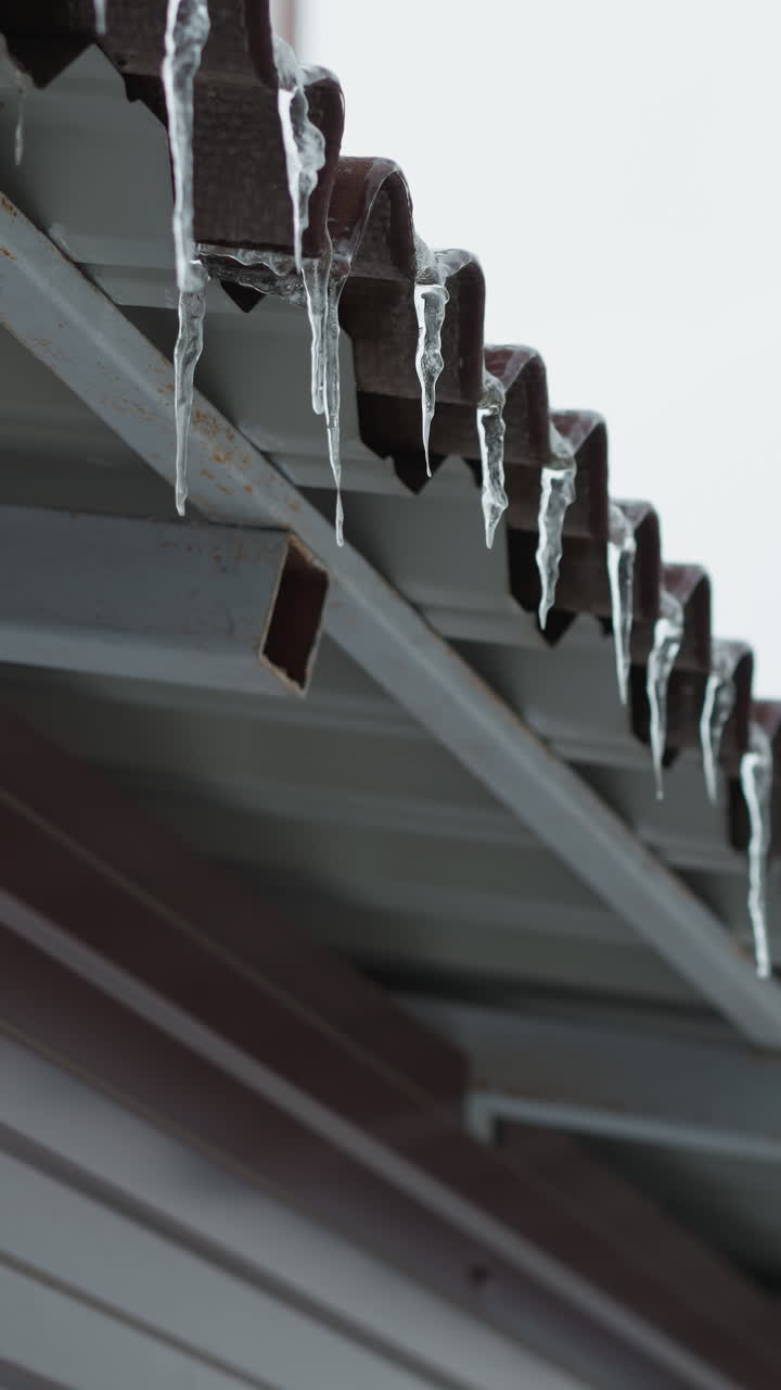 은 얼음과 매달린 얼음으로 인 지붕 가장자리의 클로즈업, 금속 구조를 따라 복잡한 얼어붙은 텍스처를 보여주는 밝은 겨울 하늘과 미니멀한 배경을 배경으로 설정