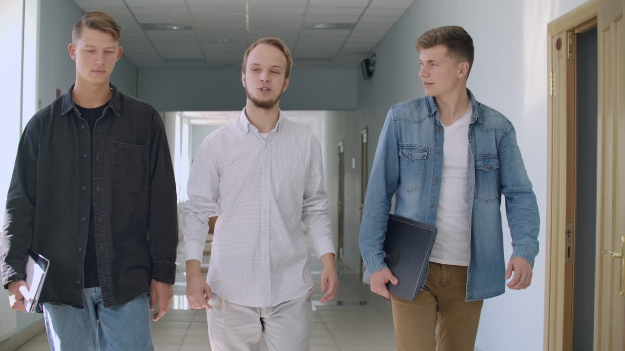 한 무리의 학생들이 대학 학교의 복도를 따라 걸어가며 대화하고 미소를 지으며 의사소통합니다. 많은 사람들이 손에 책을 들고 교육 기관의 홀에서 고 있습니다.