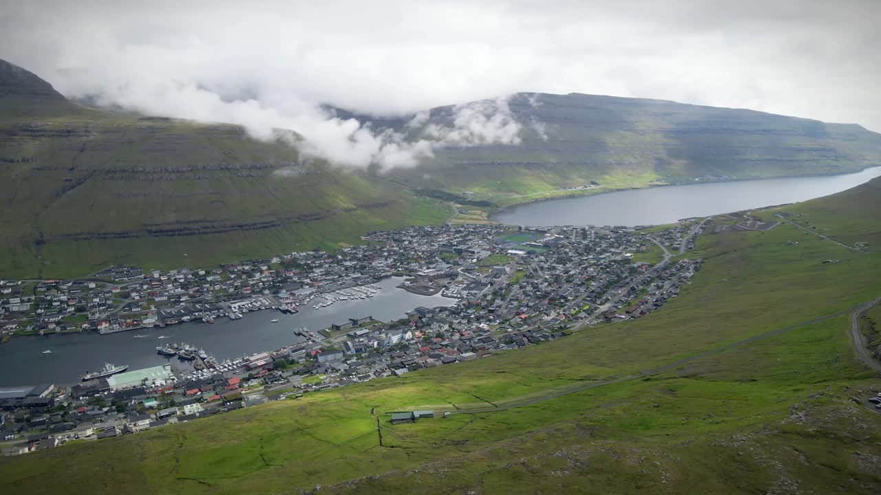 Low clouds are covering the green mountains surrounding the city of Klaksvík in the Faroe Islands, with houses and boats moored at the piers, establishing drone shot