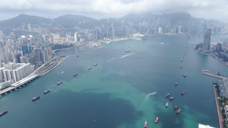 Convoy-De-Barcos-De-Pesca-Locales-Que-Causan-En-La-Bahía-Victoria-De-Hong-Kong,-Con-El-Horizonte-De-La-Ciudad-En-El-Horizonte,-Vista-Aérea