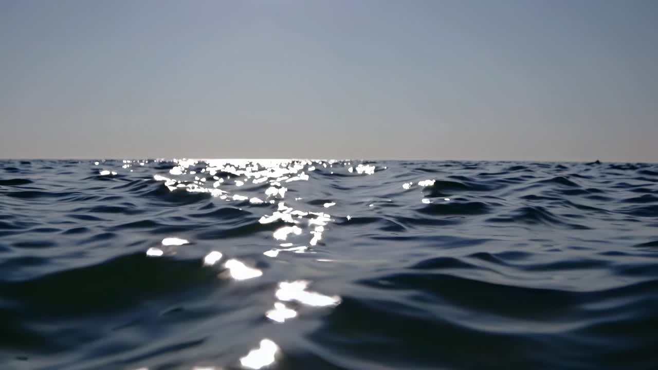 Video-opname vanuit een lage hoek van zonlicht dat weerkaatst op zachte oceaangolven, wat een serene en rustgevende sfeer creëert