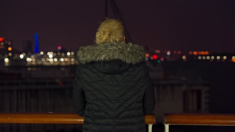 a blonde woman enjoying the beautiful view of the city at night from the balcony of the boat. view from back.