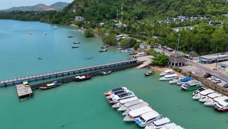drone footage captures a vibrant marina in phuket, thailand, showcasing boats, a pier, and lush surroundings under bright daylight