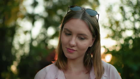 close-up of young woman looking up contemplatively with warm smile and glasses resting on her head, focusing on something in front of her, background features trees and light filtering through them