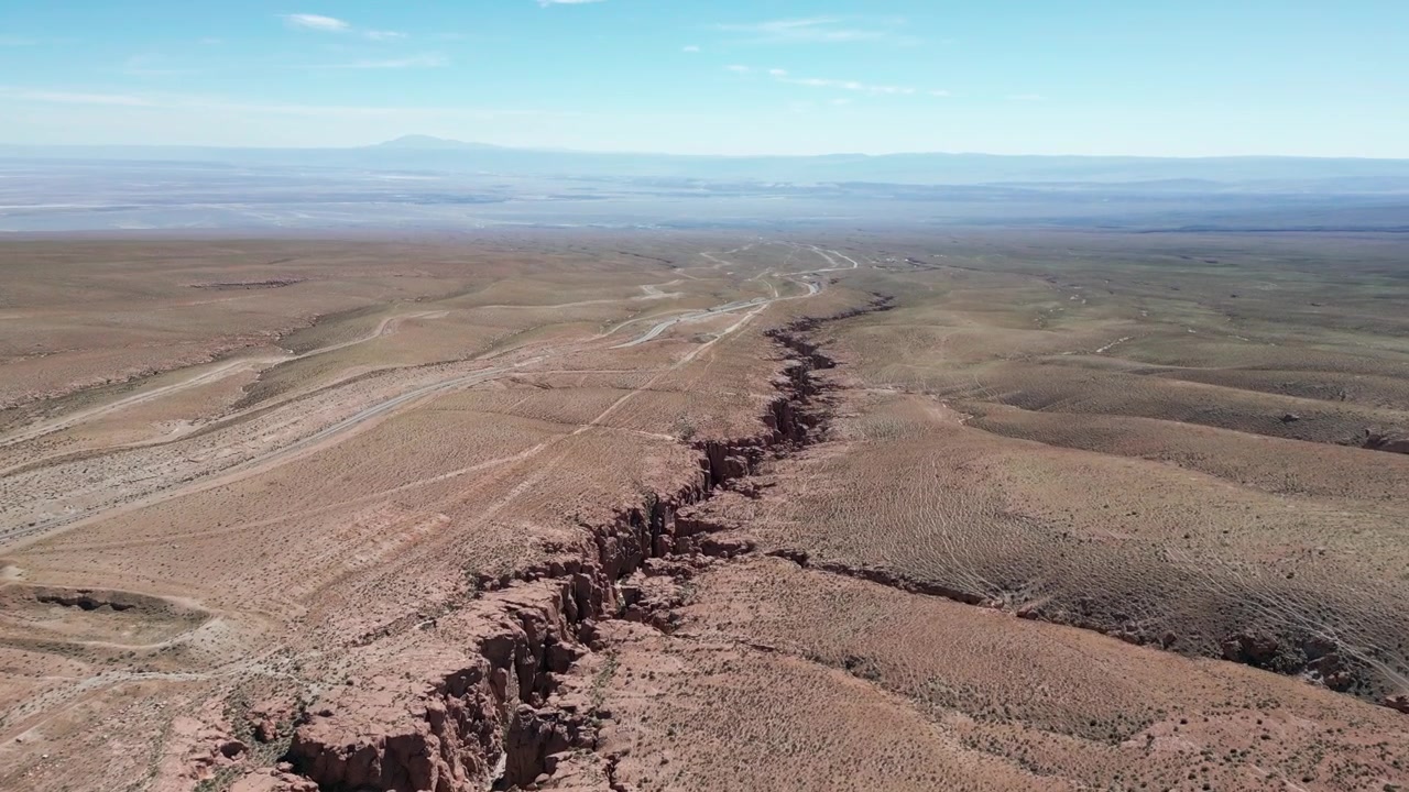 Volo in avanti ad alta quota sopra una drammatica fessura di canyon desertico che attraversa un vasto altopiano arido con un lago salato e una catena montuosa distante all'orizzonte.