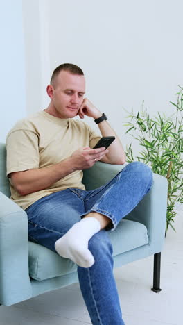 caucasian man wearing t-shirt and jeans sits in the arm-chair. man uses internet on his smartphone. vertical video.