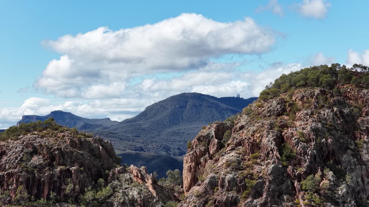 Camera slowly pans across rugged rock formations and distant mountain ranges under bright daylight, revealing dramatic Australian landscape with scattered clouds and clear blue sky