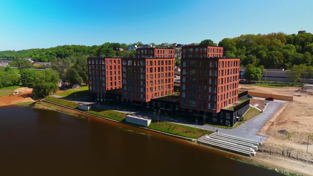 Aerial view of modern brick apartment buildings by a riverside, surrounded by greenery and clear blue skies. The scene highlights urban development and waterfront living in a serene environment.