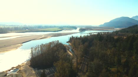 Cinematic-Scene-flying-over-the-Fraser-River-in-the-Lower-Mainland-in-British-Columbia-in-Canada,-bright-scene-with-foggy-mountains-in-the-autumn