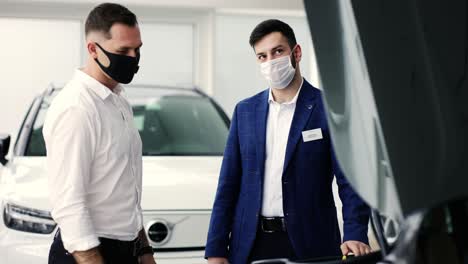 agent showing car engine under open hood to a client in auto showroom during lockdown