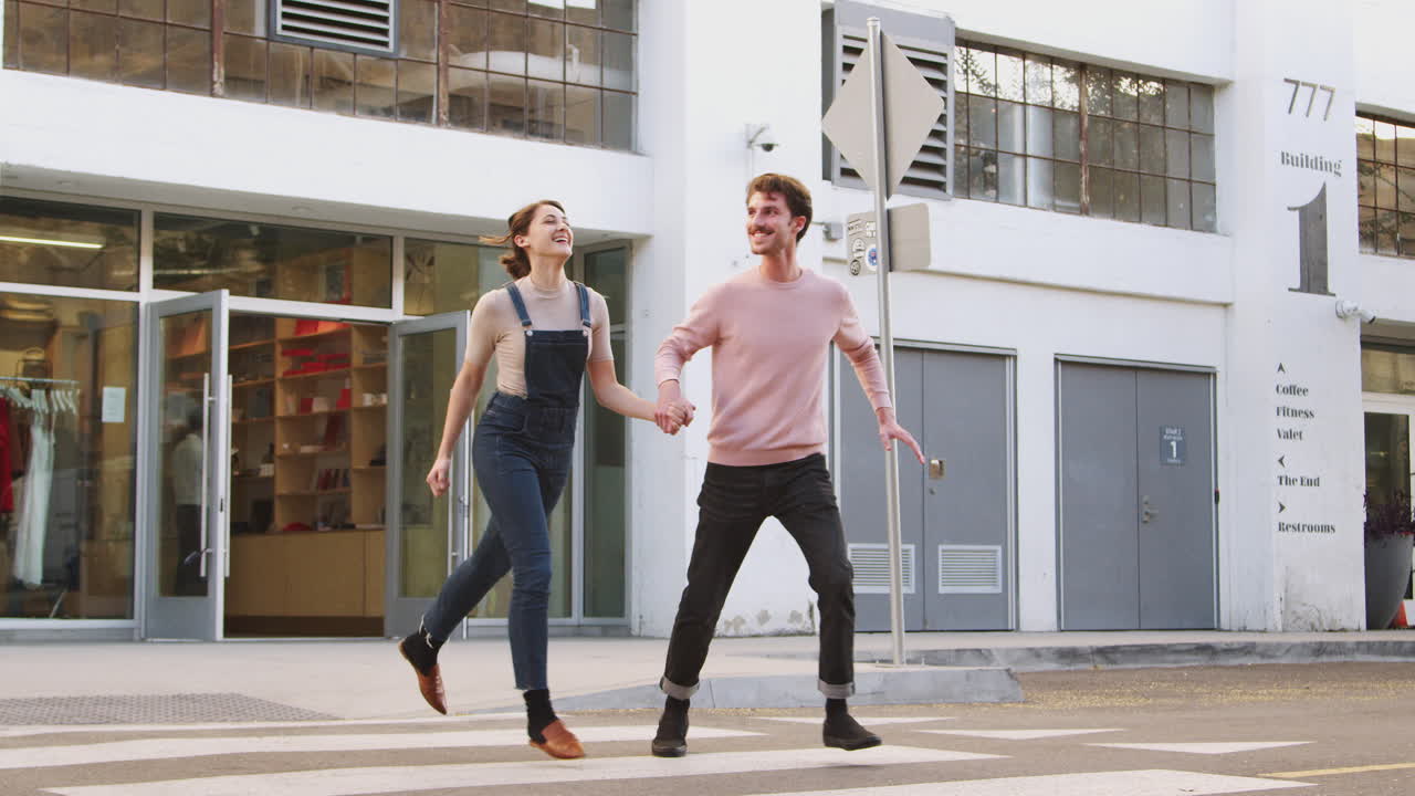 Coppia millennial hipster che attraversa la strada di corsa in città, tenendosi per mano, a figura intera.