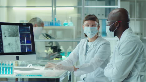 diverse colleagues in masks discussing microscopic image on computer