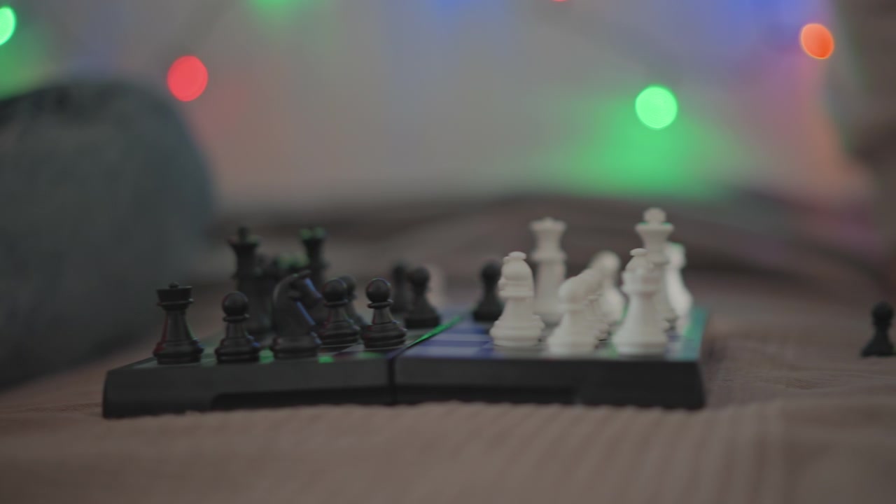 Vista ravvicinata da un'angolazione bassa mostra una scacchiera portatile durante una partita a metà gioco, con un pedone isolato, pezzi bianchi e neri raggruppati, luci festive sfocate, una superficie testurizzata, un'atmosfera interna accogliente.