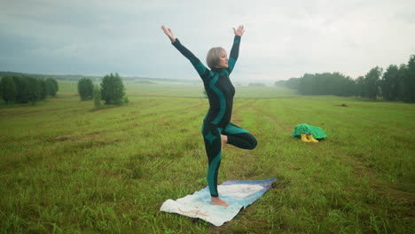 side view of woman in green and black suit practicing yoga in tree pose, lifting her hands slowly while standing on yoga mat in a vast grassy field with trees in the background under a cloudy sky