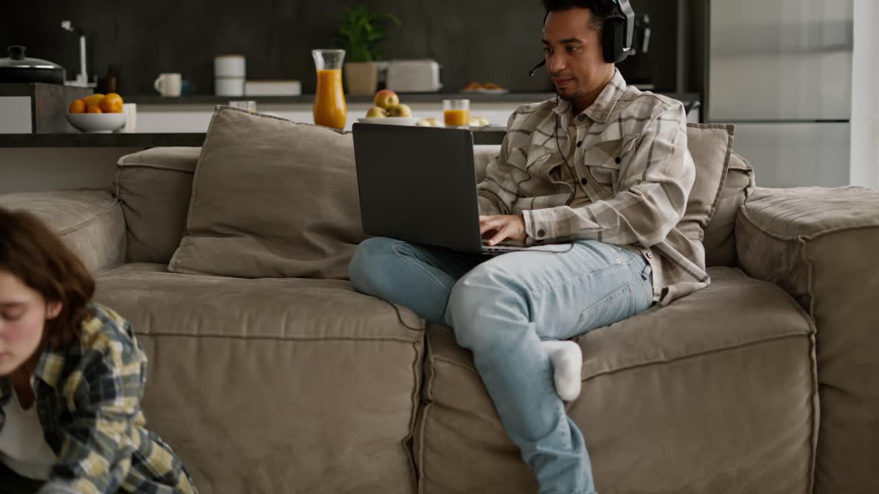 Happy young man with Black skin color in a beige checkered shirt plays computer games on his laptop while his young adult girlfriend with a bob hairstyle does yoga while sitting on the floor in a room of a modern apartment