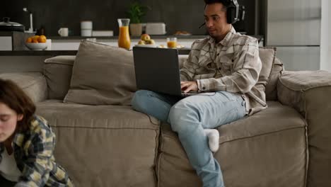 joven feliz con color de piel negra en una camisa a cuadros beige juega juegos de computadora en su portátil mientras su joven novia adulta con un peinado de bob hace yoga mientras está sentado en el suelo en una habitación de un apartamento moderno