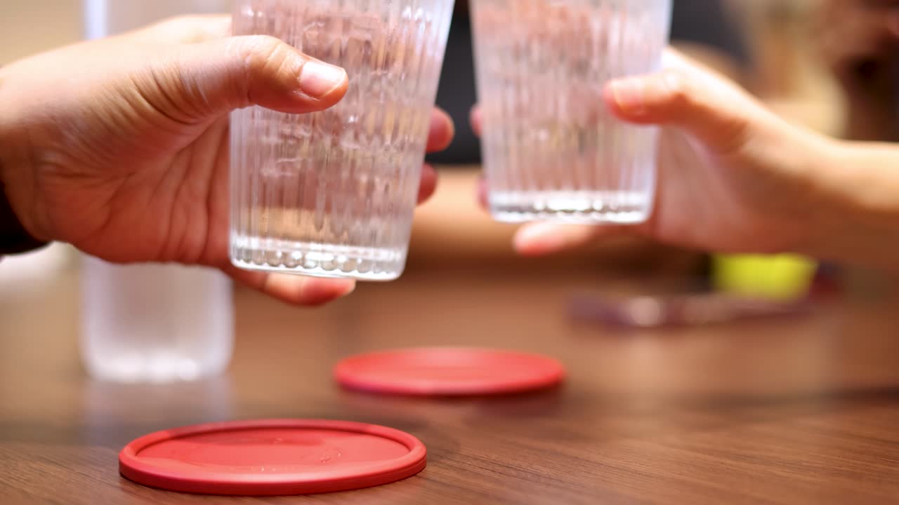 Two individuals clink glasses of water over a wooden table in a brightly lit indoor setting, with a casual, friendly atmosphere and steady camera framing