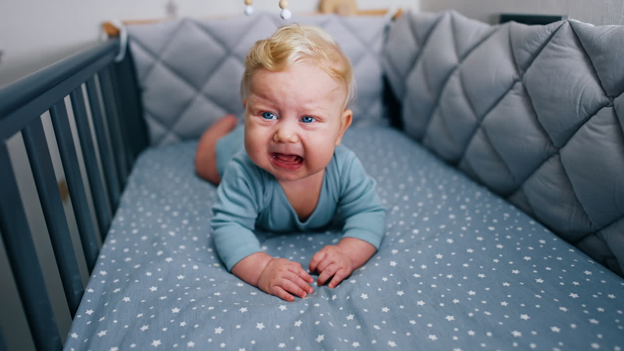 Lovely little infant boy lies in the cot and start to cry. Baby lying on the belly and crying in the crib.