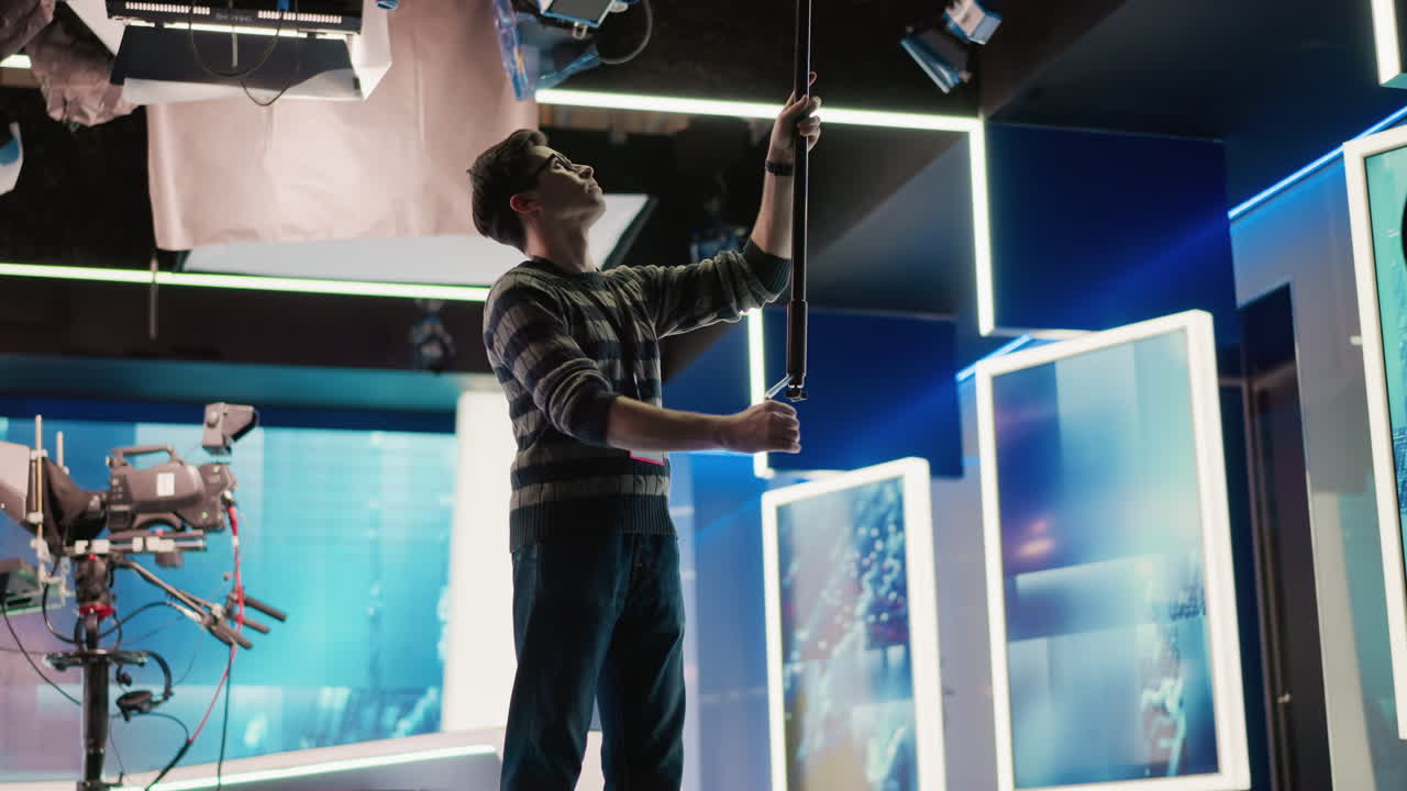 Technician adjusting boom pole in broadcast studio, preparing for shot. Modern media production setup with equipment and studio lighting visible, showcasing professional production environment