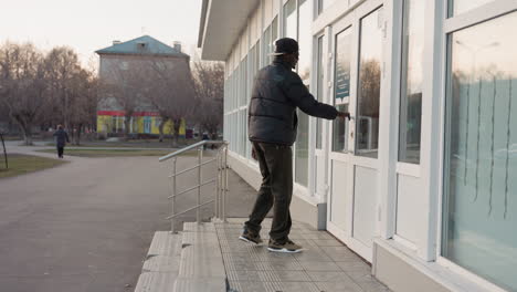 side view of young man walking along sidewalk past building with glass windows during dusk in urban neighborhood with pedestrians in background and bare trees