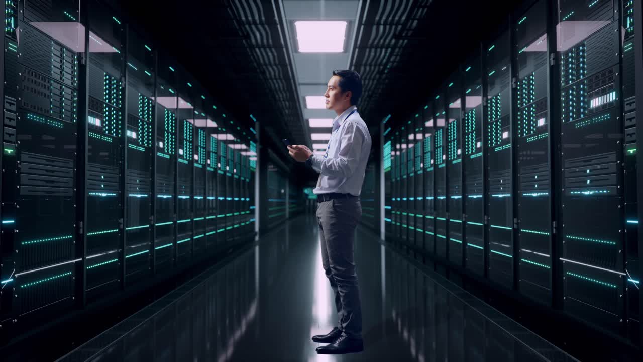Fuldkropsbillede fra siden af en asiatisk mandlig medarbejder, der står med sin telefon i et serverrum i et datacenter. Han observerer ved at se sig omkring, før han koncentrerer sig om sin smartphone.
