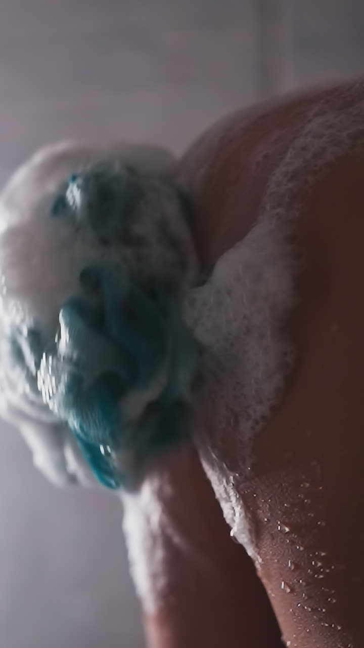 Woman rubs skin with loofah in shower cabin closeup. Lady washes body with soft foamy sponge near falling water drops. Skincare and massage tool