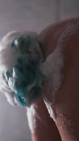 woman rubs skin with loofah in shower cabin closeup. lady washes body with soft foamy sponge near falling water drops. skincare and massage tool