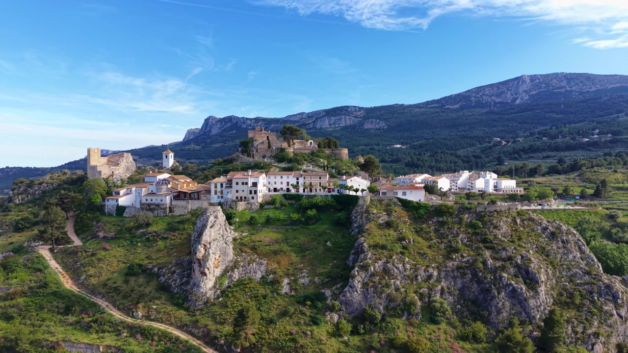 Castello e villaggio medievale di Guadalest arroccato su una collina rocciosa in Spagna, circondato da montagne e valli verdi sotto un cielo blu con nuvole leggere