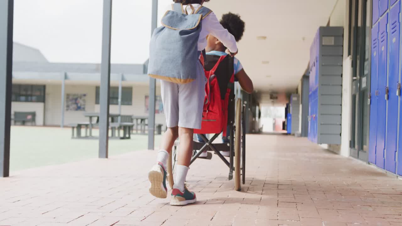 vista trasera de video de dos niños diversos, niño empujando a una niña en silla de ruedas en la escuela, con espacio para copiar