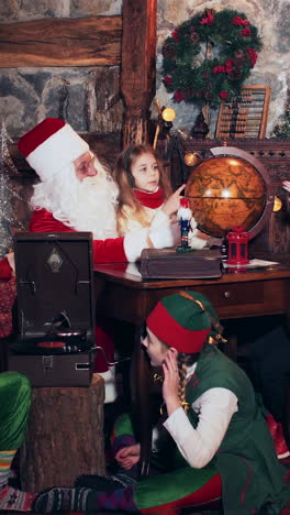 a boy and a girl, together with santa, are exploring countries on an old globe. christmas elves near the fireplace listening to a retro gramophone. vertical video