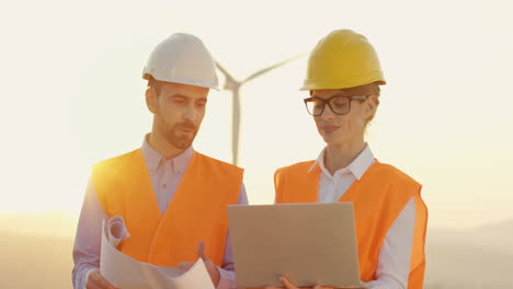 ingenieros caucásicos masculinos y femeninos con casco y uniforme usando laptop y mirando planos en la estación eólica de energía renovable