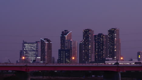 Tráfico-Conduciendo-En-Una-Autopista-Elevada-Con-Edificios-De-Gran-Altura-Iluminados-En-El-área-Metropolitana-De-Tokio,-Japón-Al-Atardecer