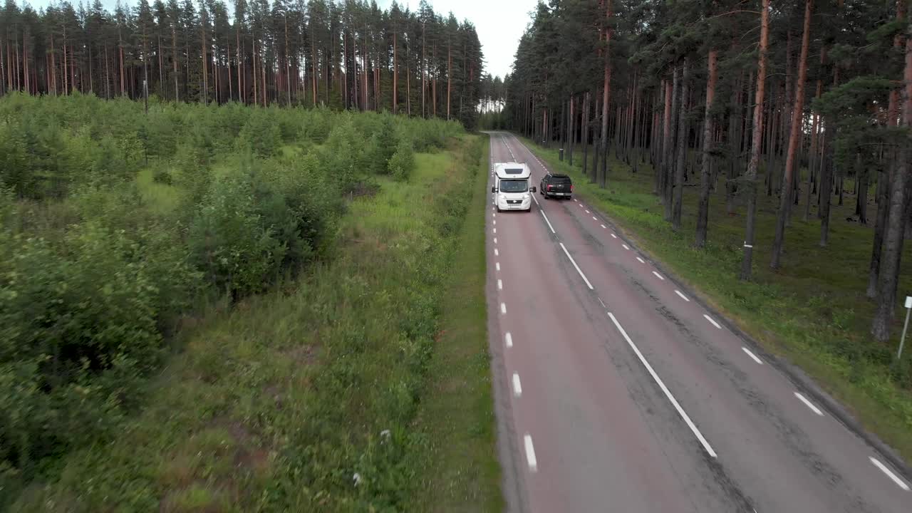 Ripresa aerea all'indietro che segue un camper bianco mentre attraversa una strada forestale a livello del suolo, circondato da alti pini, in una foresta boreale nordica