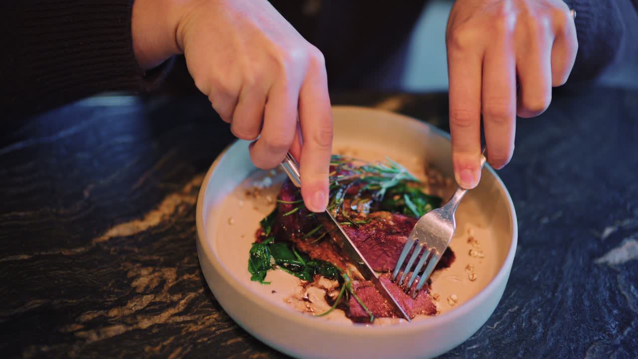 Close up of a woman's hands cutting and eating a plated steak with fork and knife